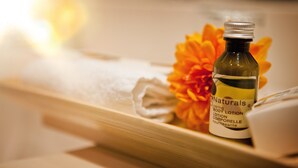 Una botella de loción corporal Naturals sobre una bandeja de madera con una toalla blanca doblada y una flor naranja brillante.