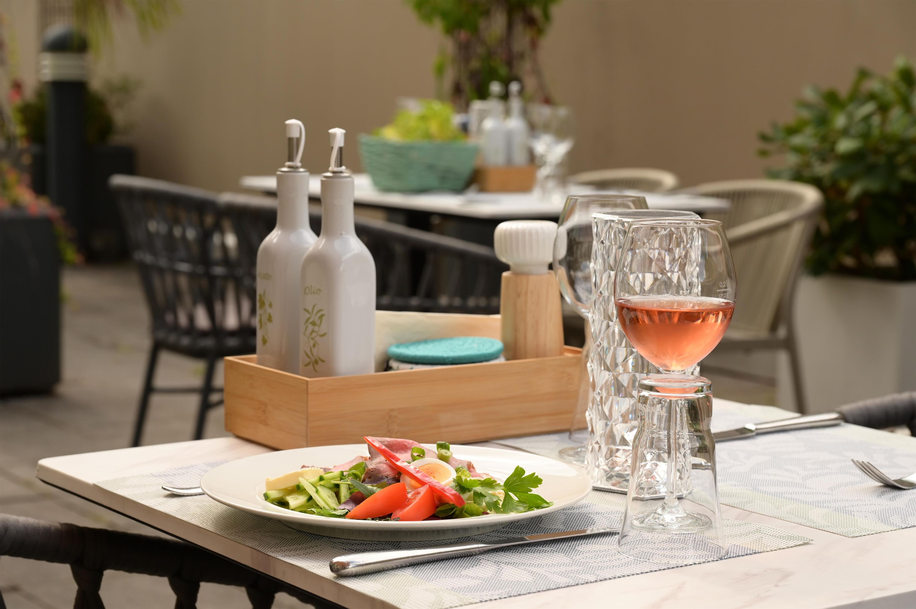 Un espacio de comedor al aire libre con una mesa preparada para dos personas, que incluye una ensalada, una copa de vino rosado y botellas decorativas.