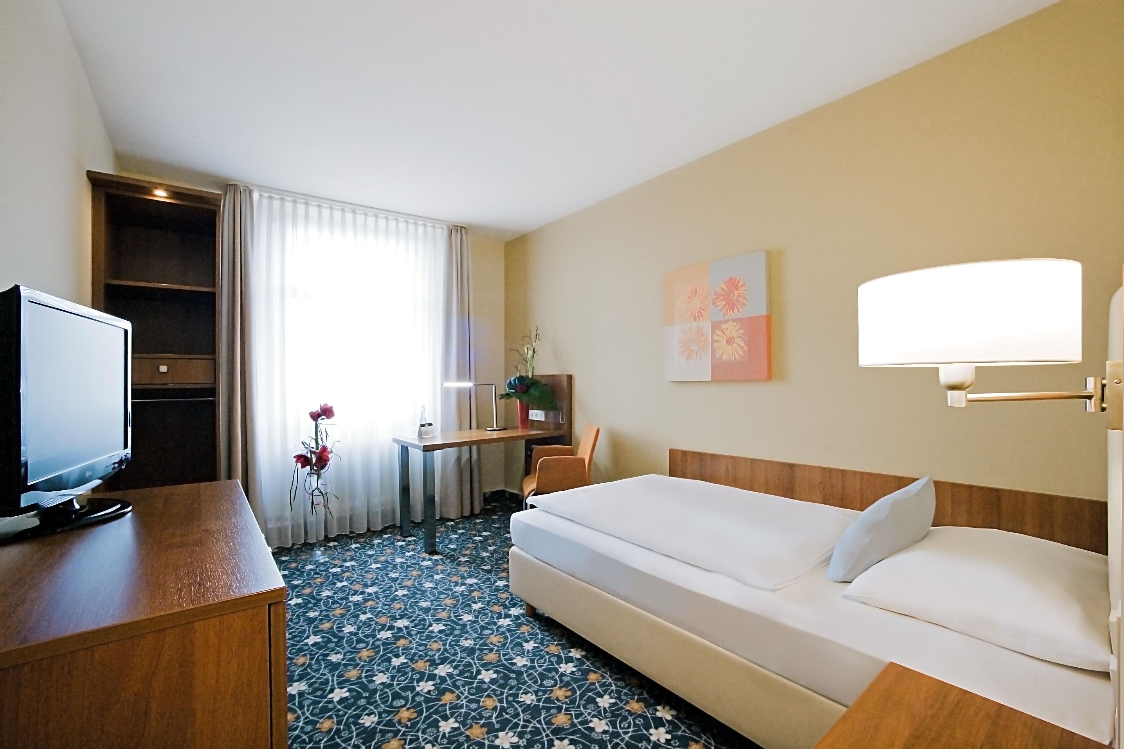 A hotel room with a bed, a desk with a computer, a television, and a window with curtains.