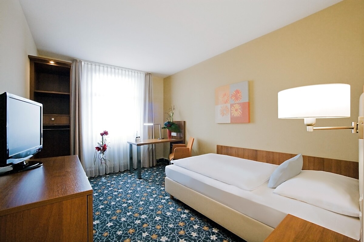 Una habitación de hotel con una cama, un escritorio con computadora, televisor y ventana con cortinas.