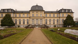 A large, historic building with symmetrical architecture, a central dome, and a formal garden with pathways and benches.
