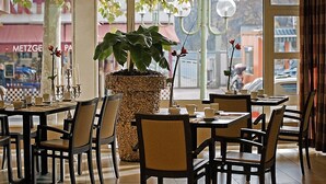 A café with tables and chairs, a large woven planter with a plant, and a view of the street outside.