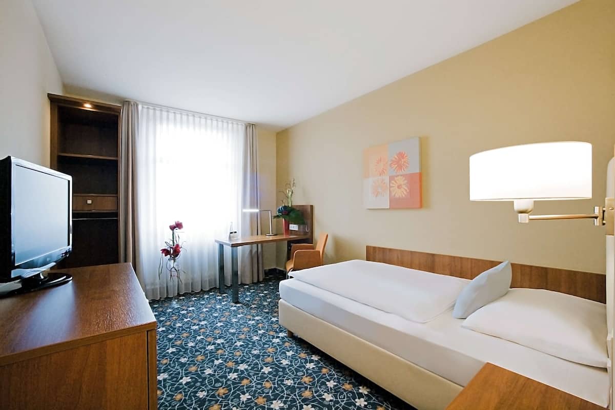 Une chambre d’hôtel avec un lit, un bureau avec une télévision, une petite table avec un vase de fleurs et une fenêtre avec des rideaux.