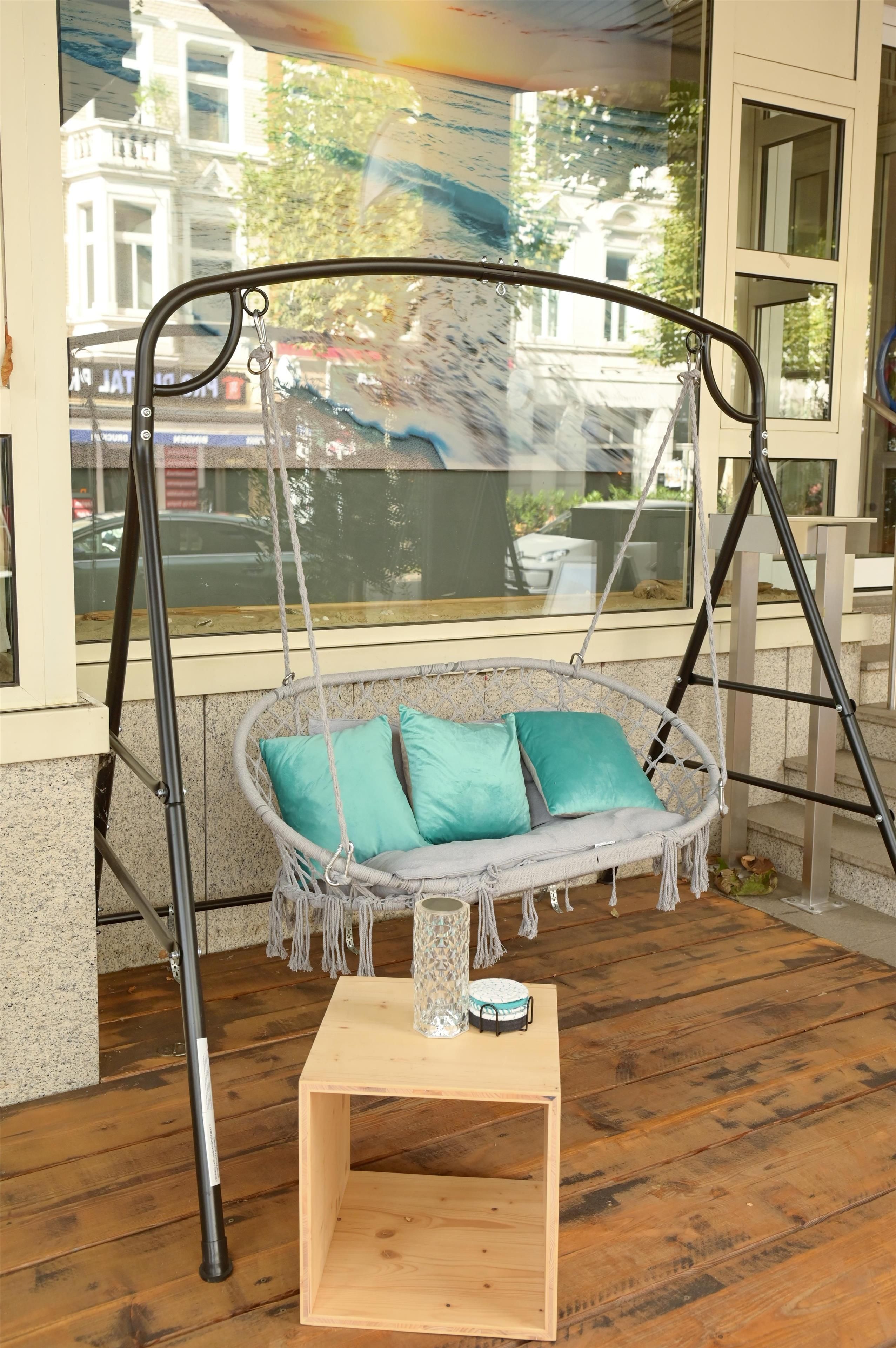 A wooden swing chair with teal cushions, a small table, and a window with a view of a building and parked cars.