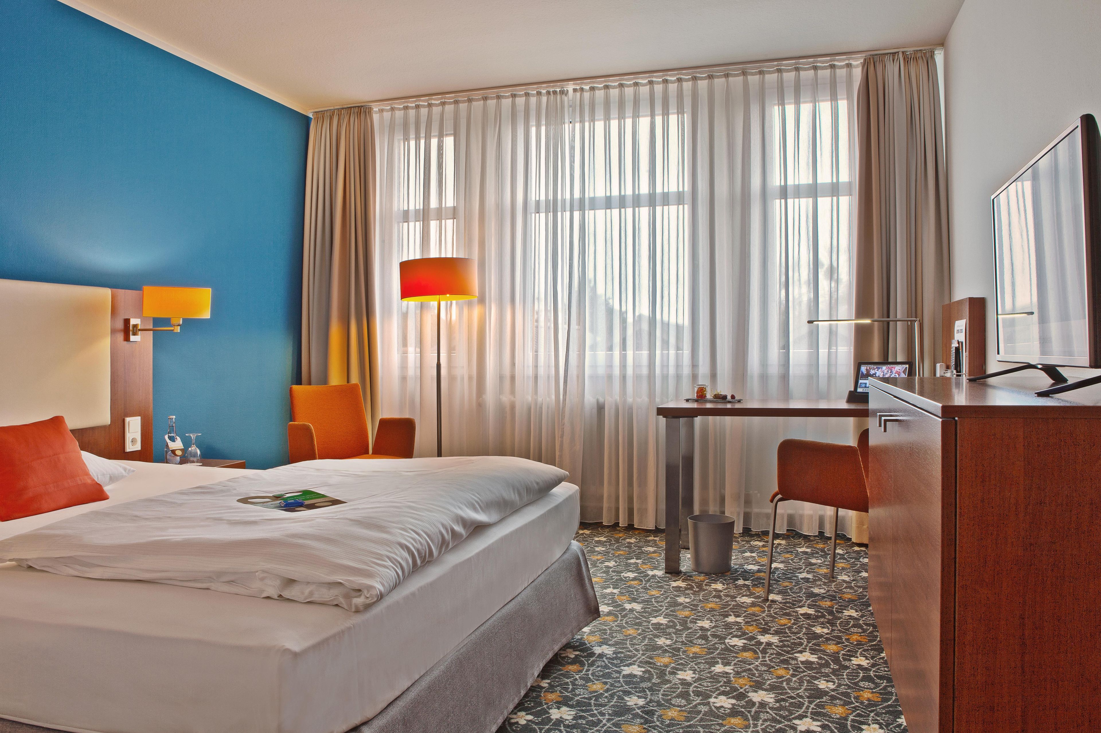 A hotel room with a bed, a desk, a chair, a lamp, and a television.