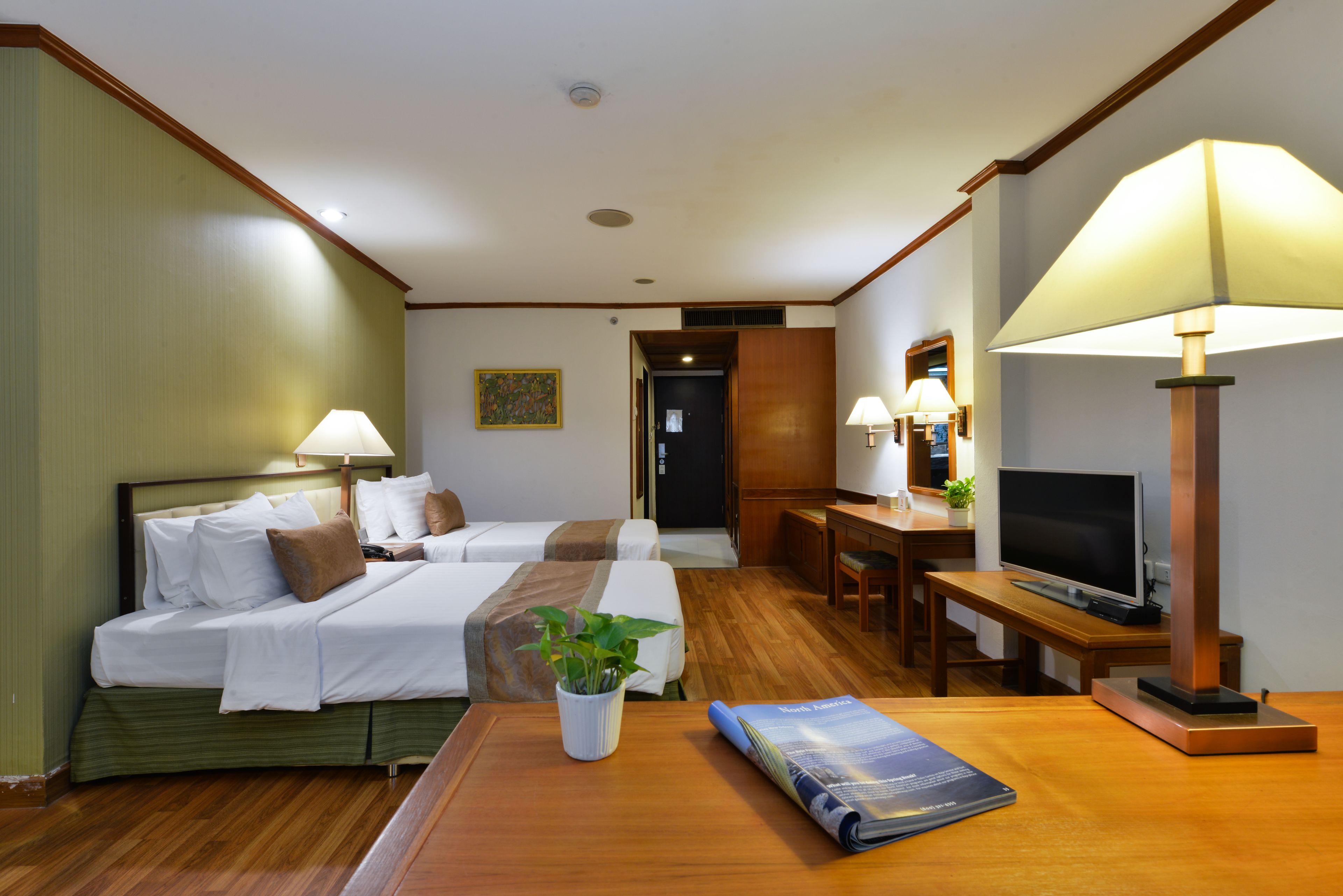 A hotel room with two beds, a desk with a television, a lamp, and a plant.