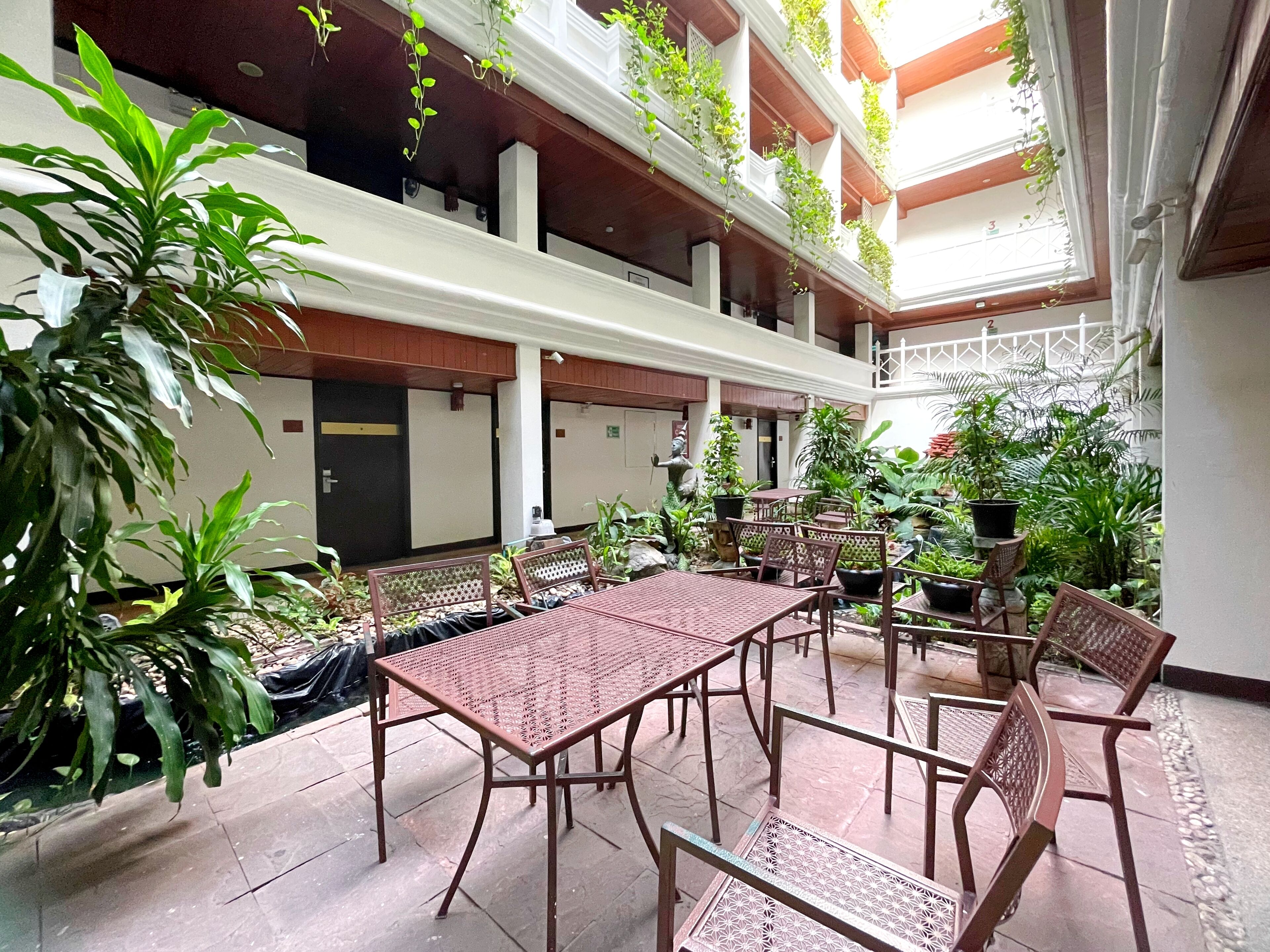 Une cour intérieure spacieuse, dotée de nombreuses tables et chaises, et entourée d’une végétation luxuriante et de plantes en pot.