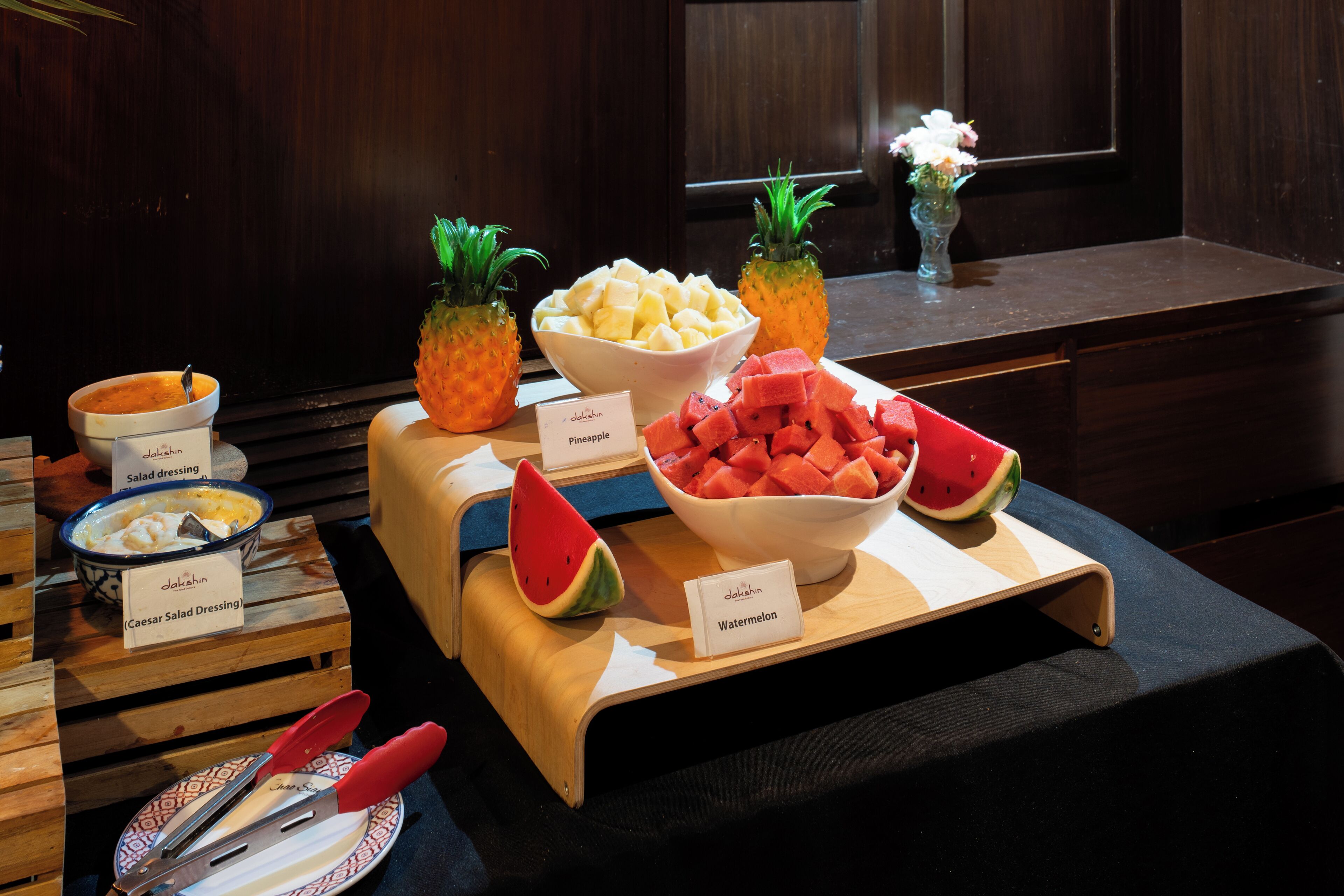Une table de buffet avec des bols contenant de la vinaigrette César, des ananas, des fruits coupés en dés et des pastèques.
