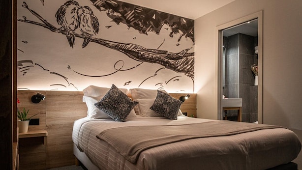 Un dormitorio con una cama grande, dos almohadas decorativas, una mesita de noche con una planta y un mural en la pared.