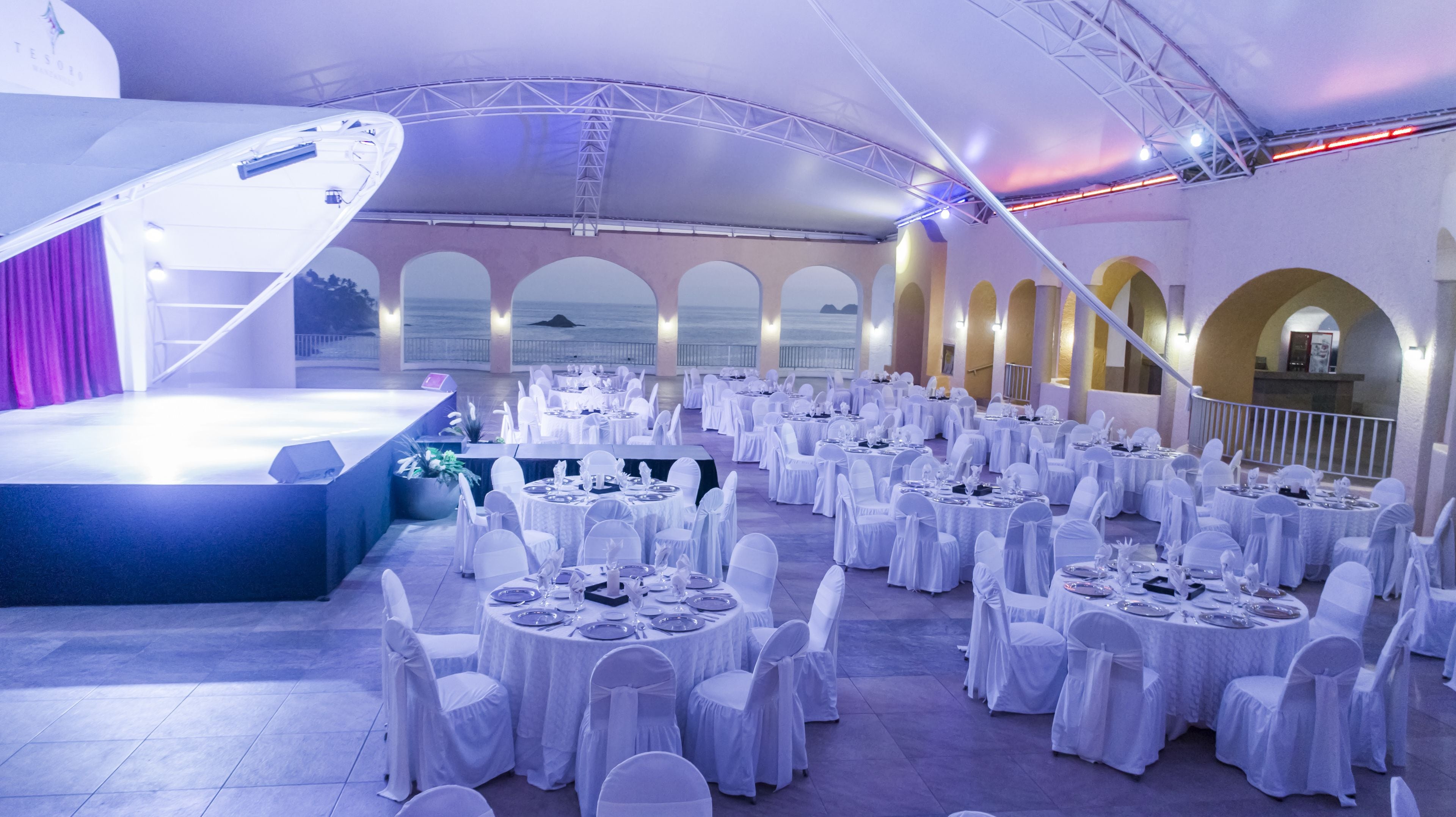 Un salón de banquetes con mesas redondas y sillas blancas, un escenario con fondo azul y ventanas arqueadas que dan a un cuerpo de agua.