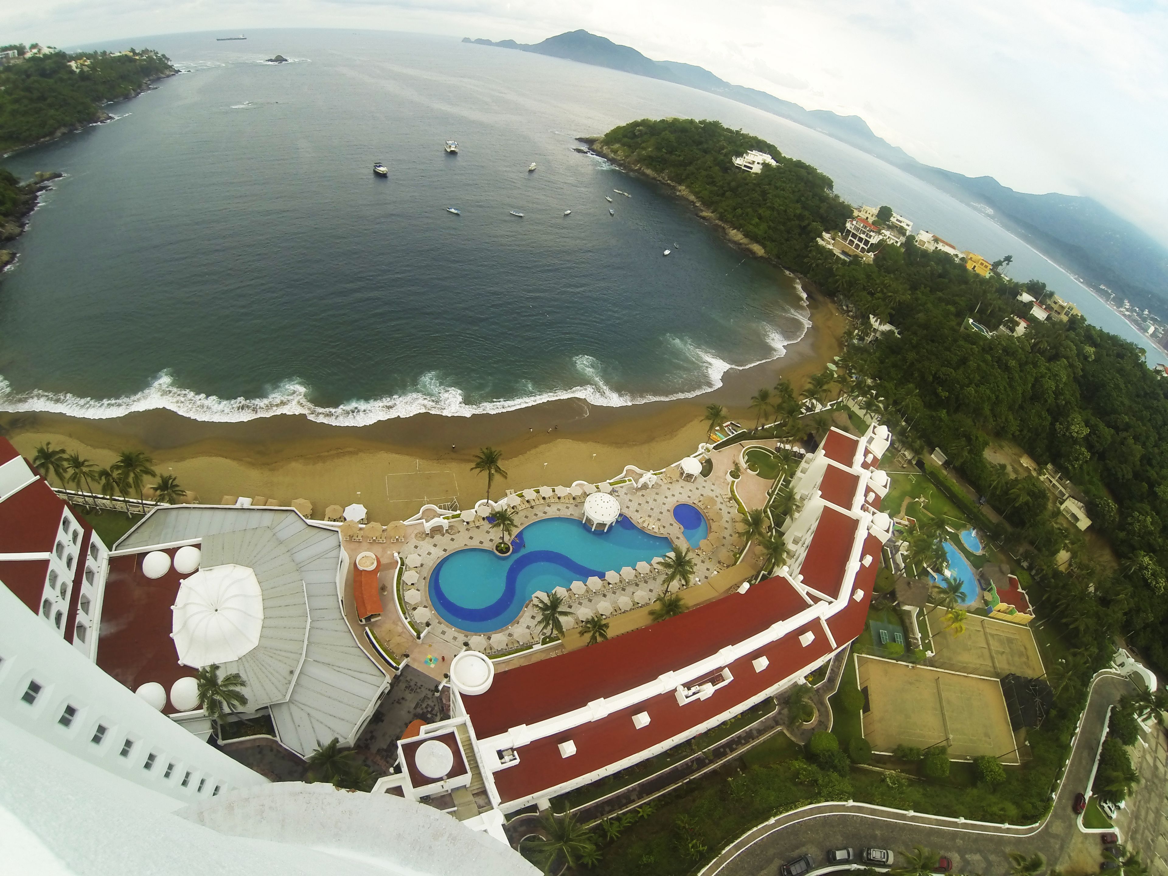 An aerial view of a coastal resort with a beach, swimming pools, and a parking area.