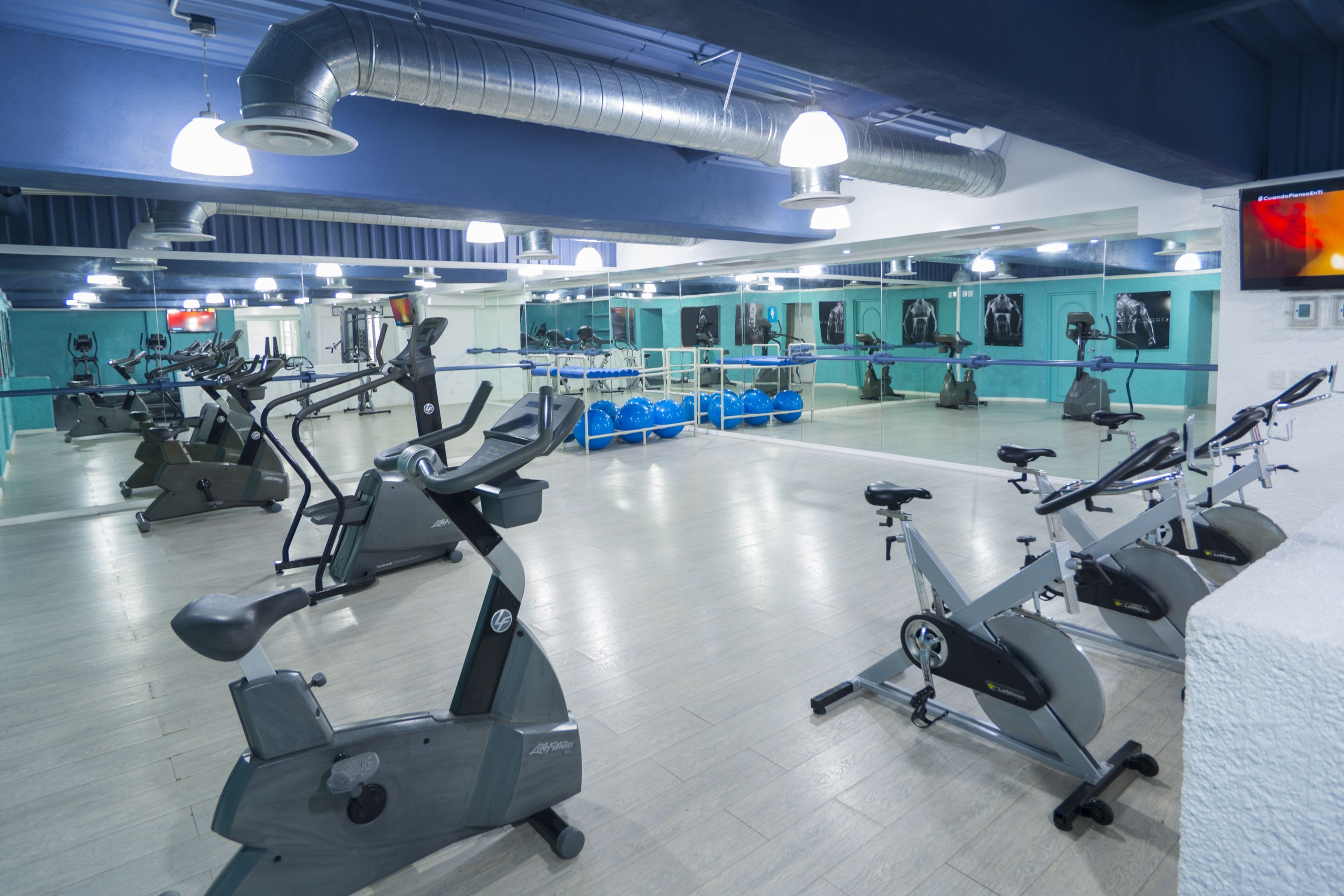 Un gimnasio con varias bicicletas estáticas y máquinas de cardio, un espejo grande y una pantalla mostrando contenido.