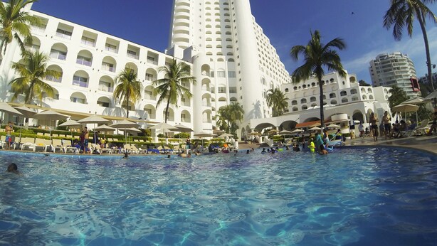 Un hotel con piscina, palmeras y un edificio grande con varios balcones.