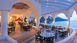 Un espacio exterior para comer con mesas y sillas, un paseo cubierto y vistas al mar.