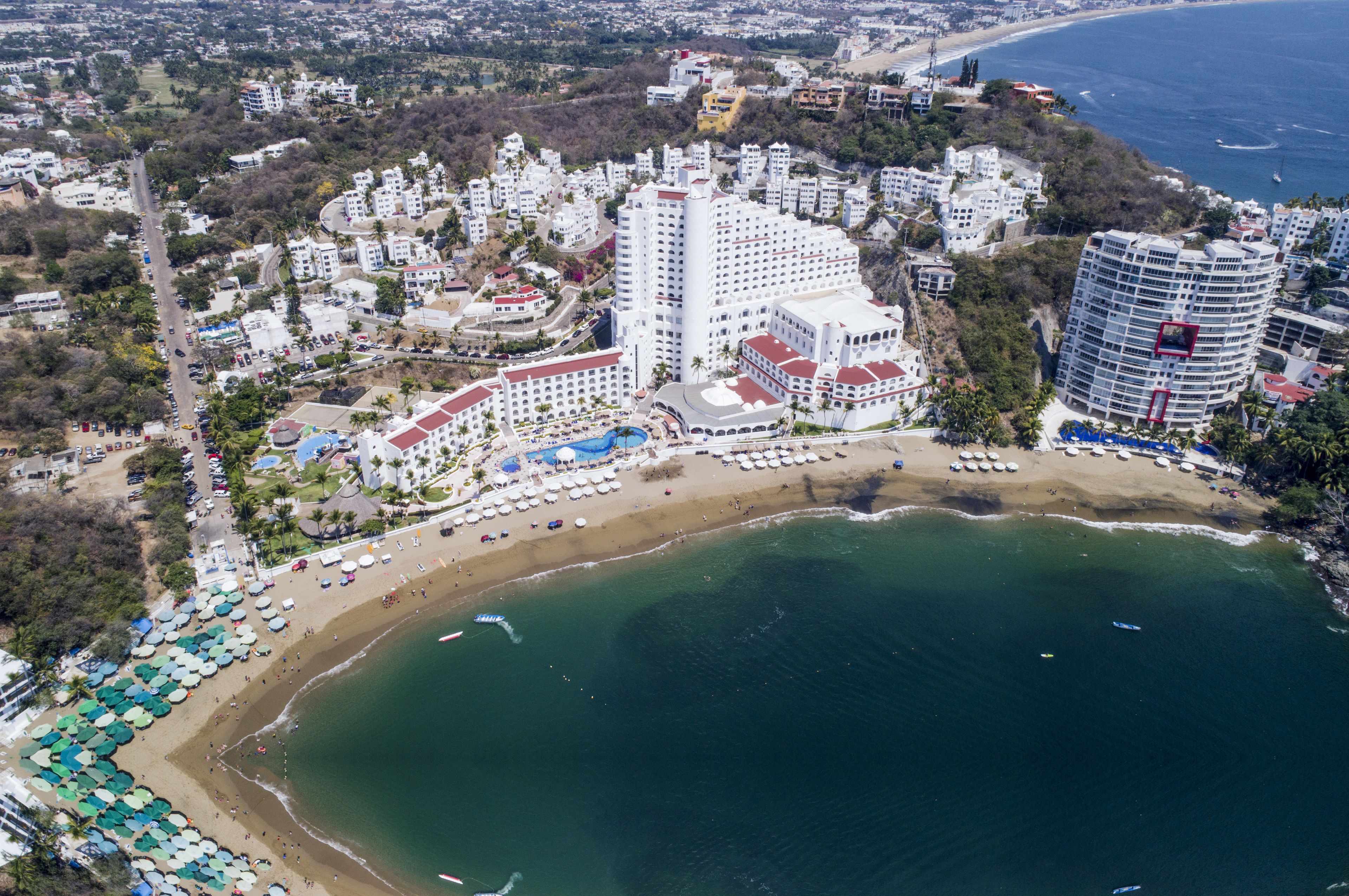 Vista aérea de un complejo turístico costero con varios hoteles de gran altura, una playa de arena y una vista despejada del océano.