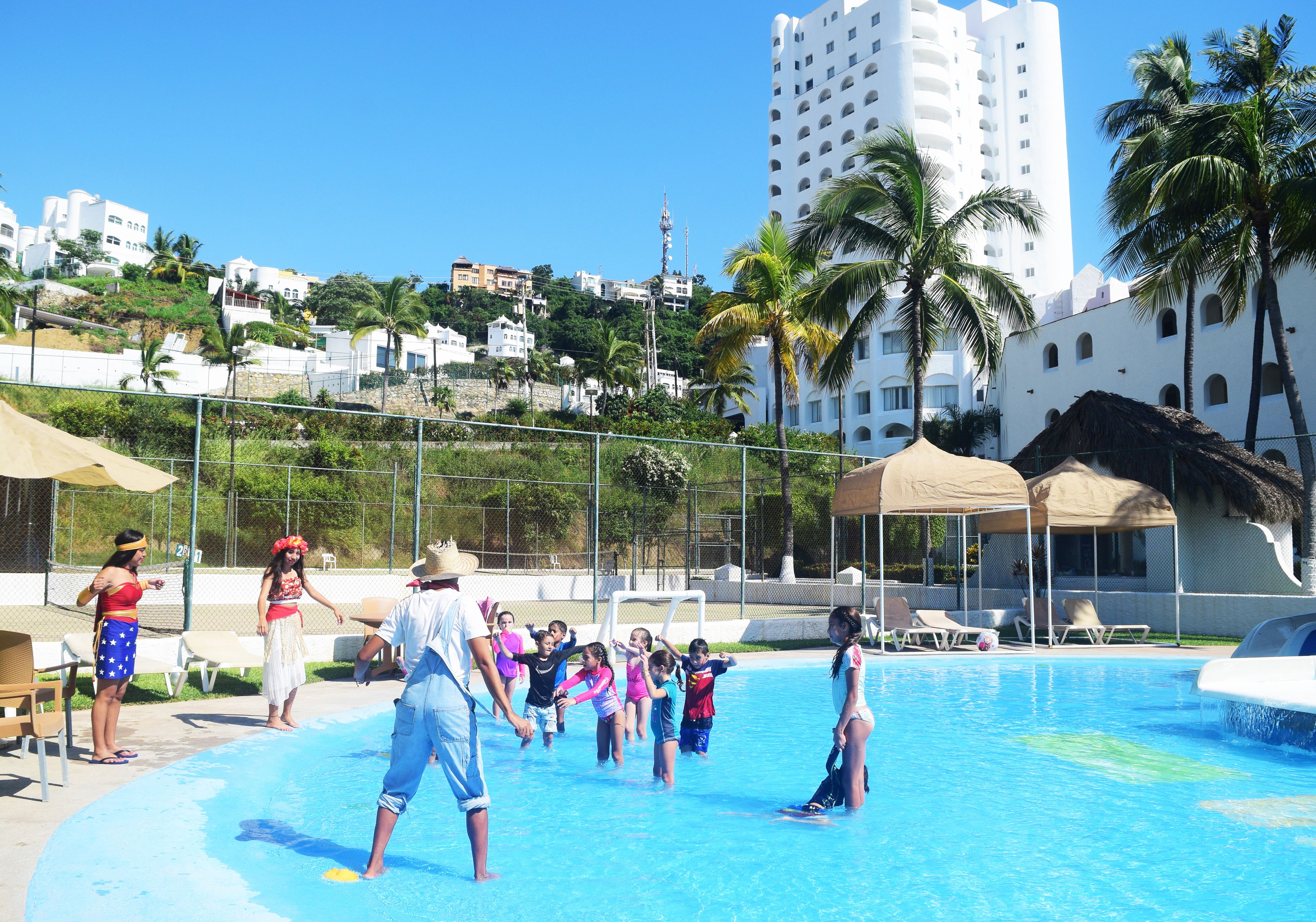 A swimming pool area with a clear sky, palm trees, and buildings in the background.
