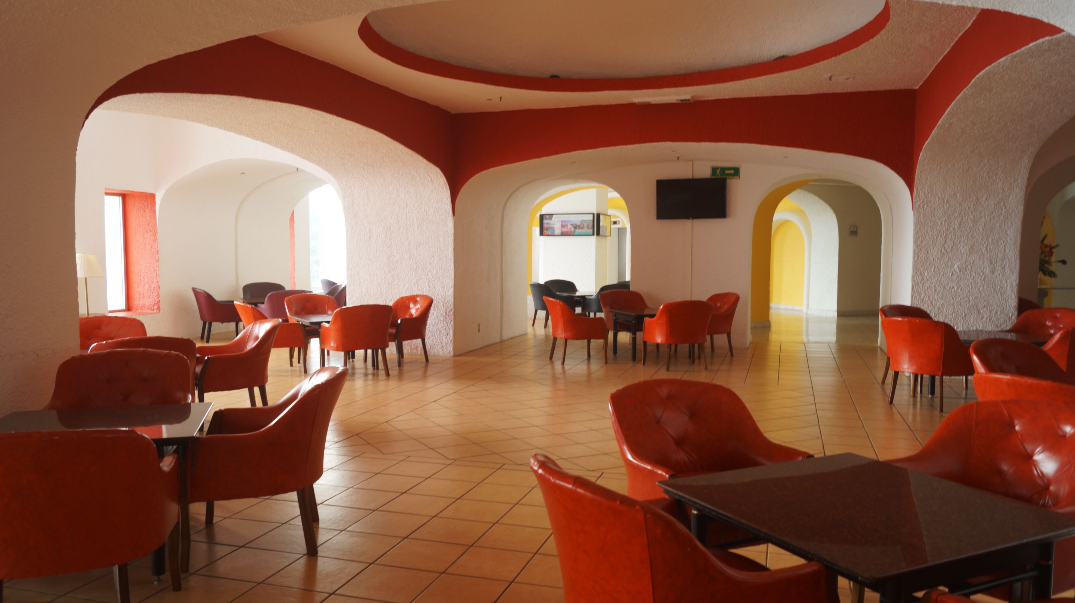 Un comedor con sillas y mesas rojas, paredes blancas y suelo de baldosas.