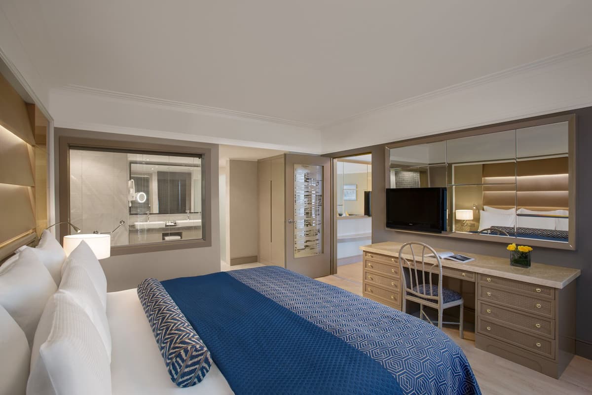 Quarto de hotel moderno com uma cama grande, escrivaninha, cadeira, televisão e banheiro com espelho e pia.