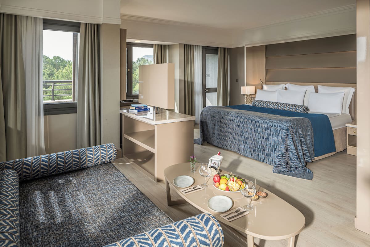 Quarto de hotel com cama, sofá, mesa de jantar com pratos e copos, e uma pequena mesa com uma fruteira.
