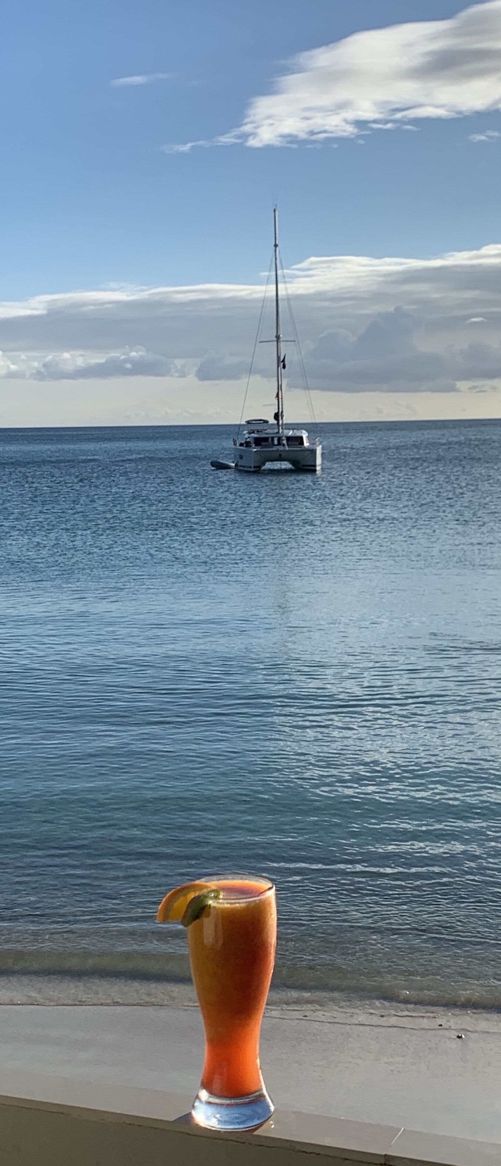 A glass of drink on a ledge with a view of a calm sea and a sailboat in the distance.