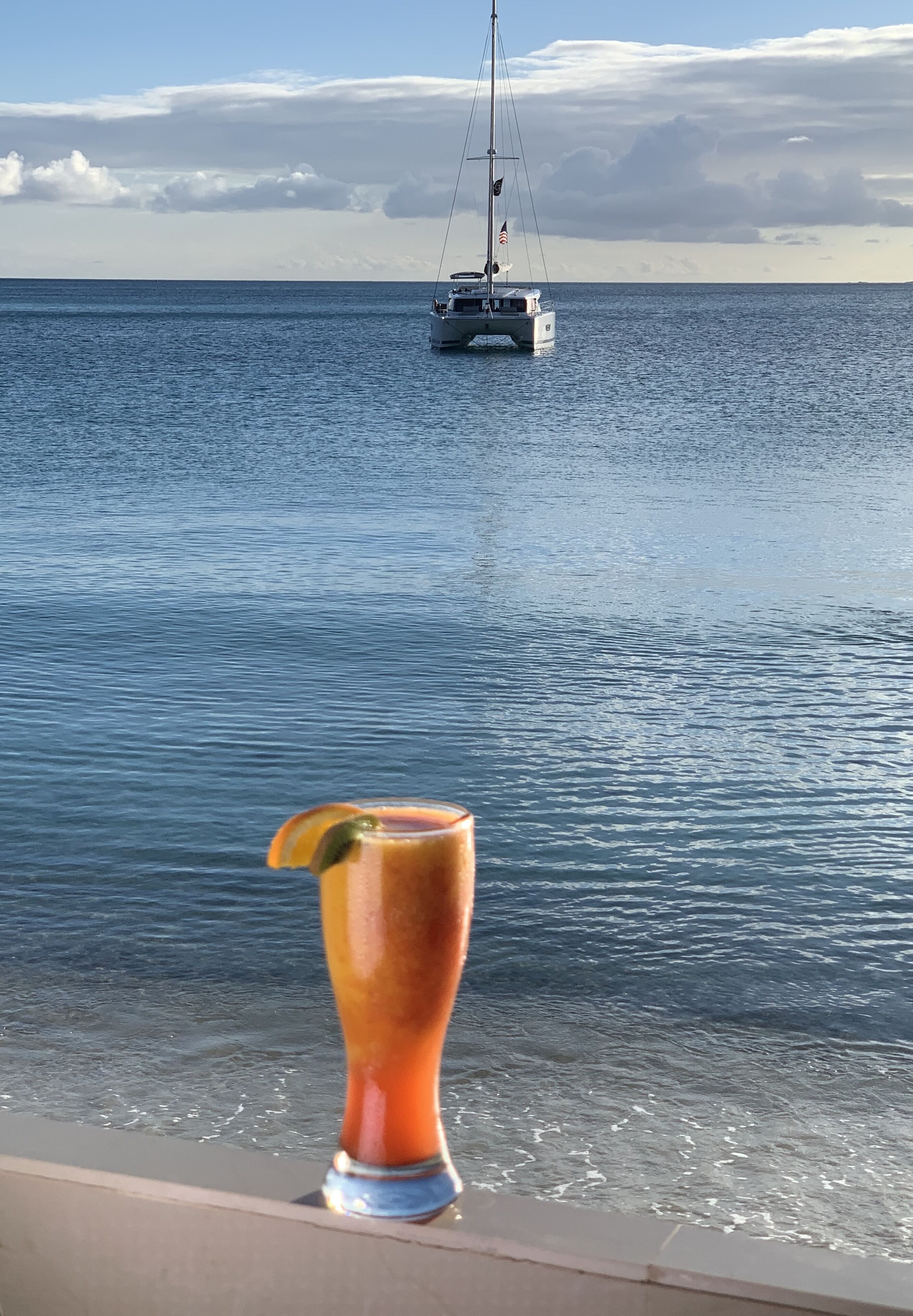 Un verre à la main, sur un balcon donnant sur une mer calme, avec un voilier à l'horizon.