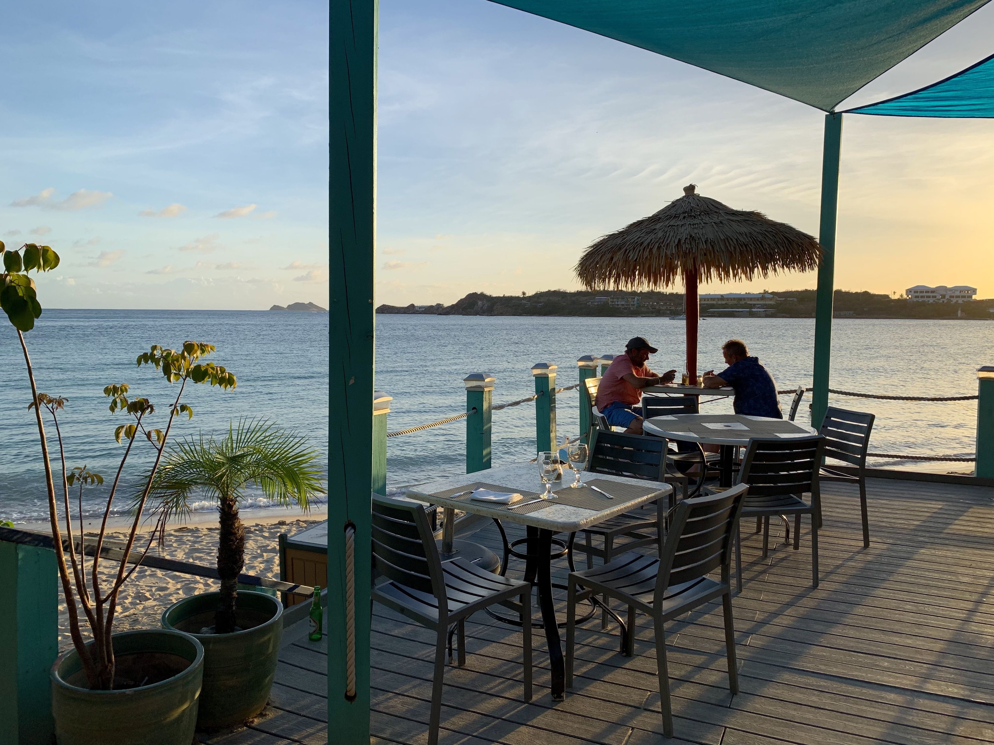 A seaside restaurant with outdoor seating, a thatched umbrella, and a view of the ocean.