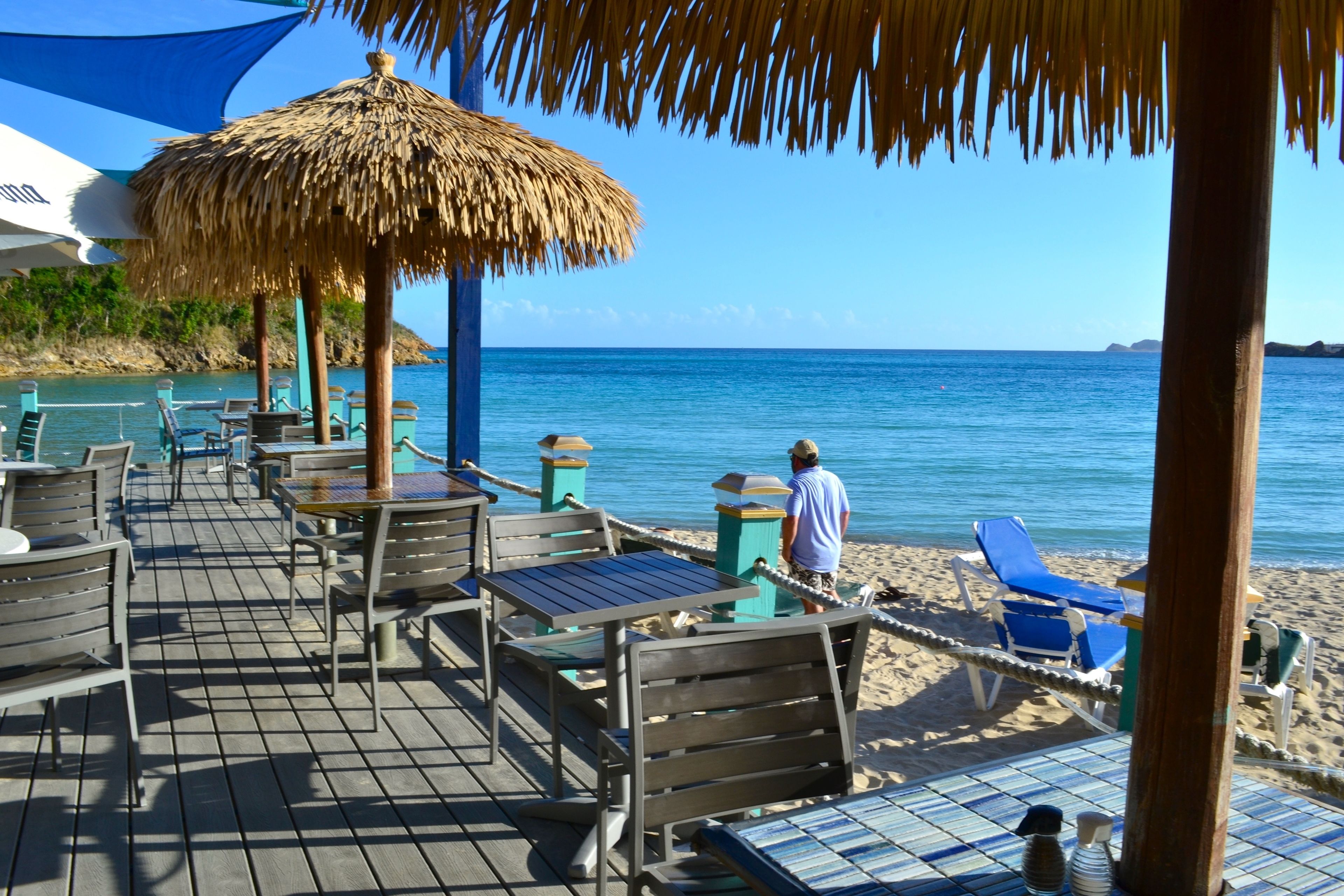 A beachside patio with wooden tables and chairs, a thatched umbrella, and a view of the ocean.