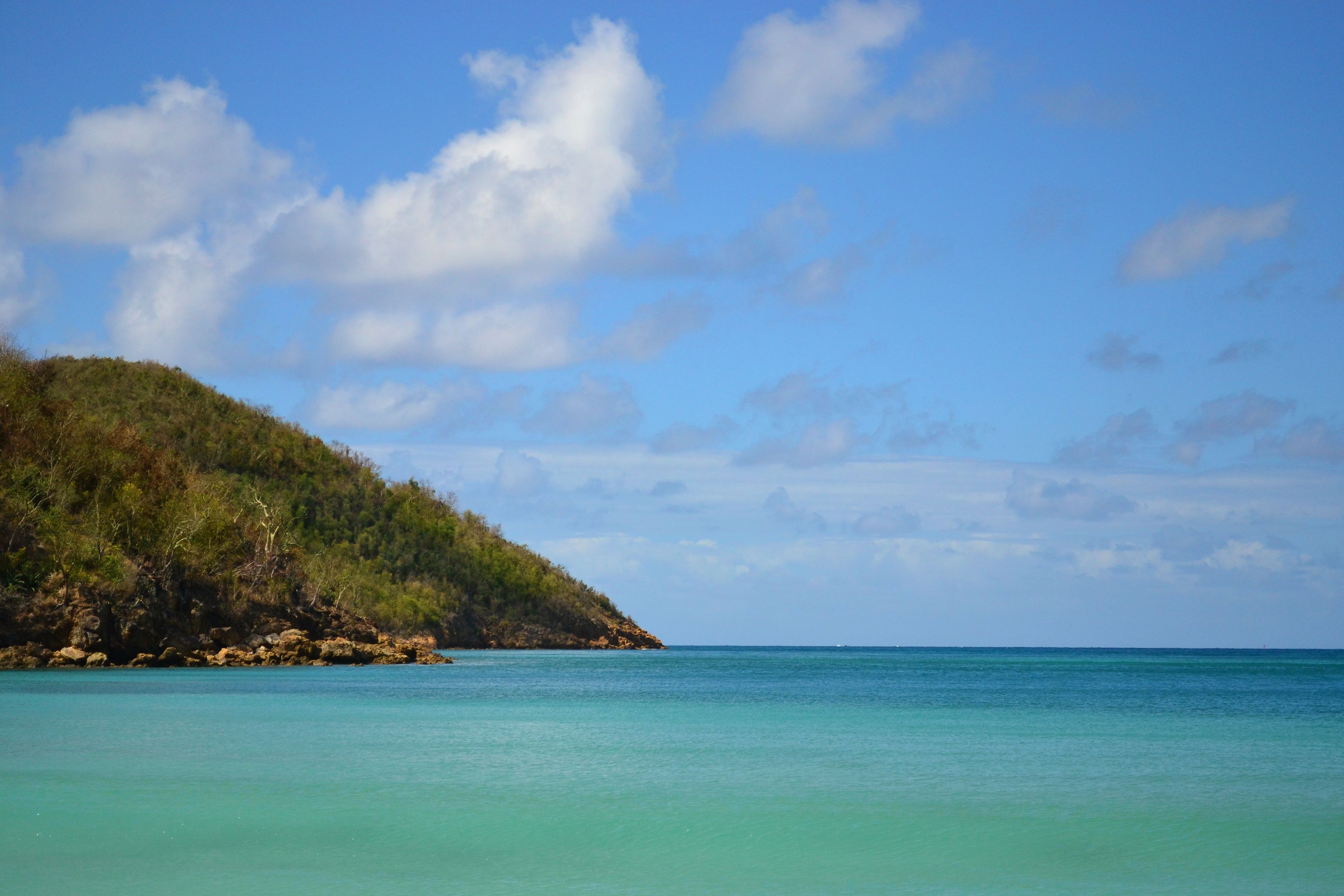 A coastal landscape with clear turquoise waters and a green hill with trees.