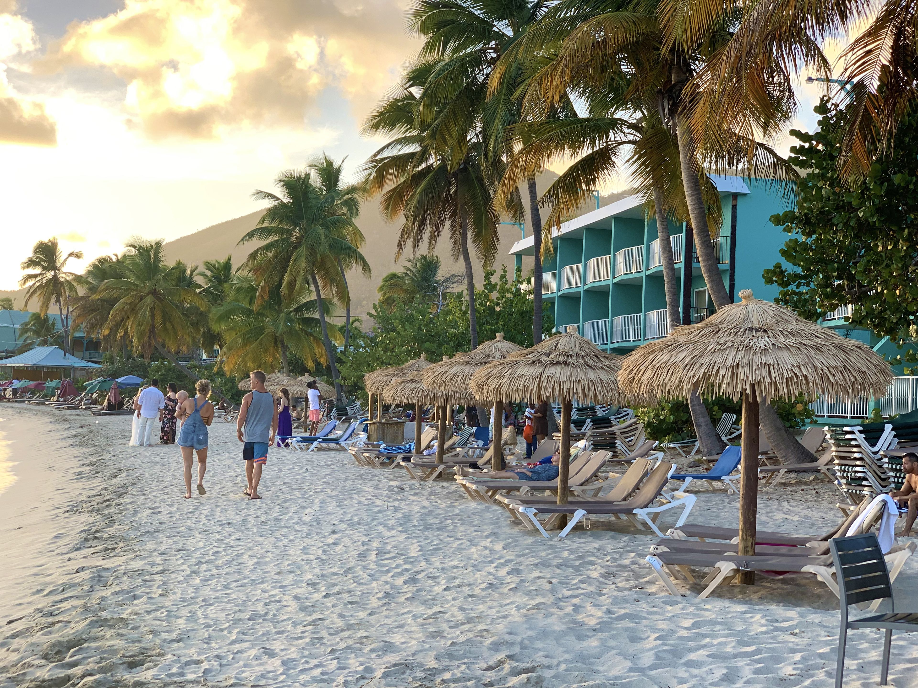 A beach with lounge chairs and thatched umbrellas, palm trees, and a building with balconies.