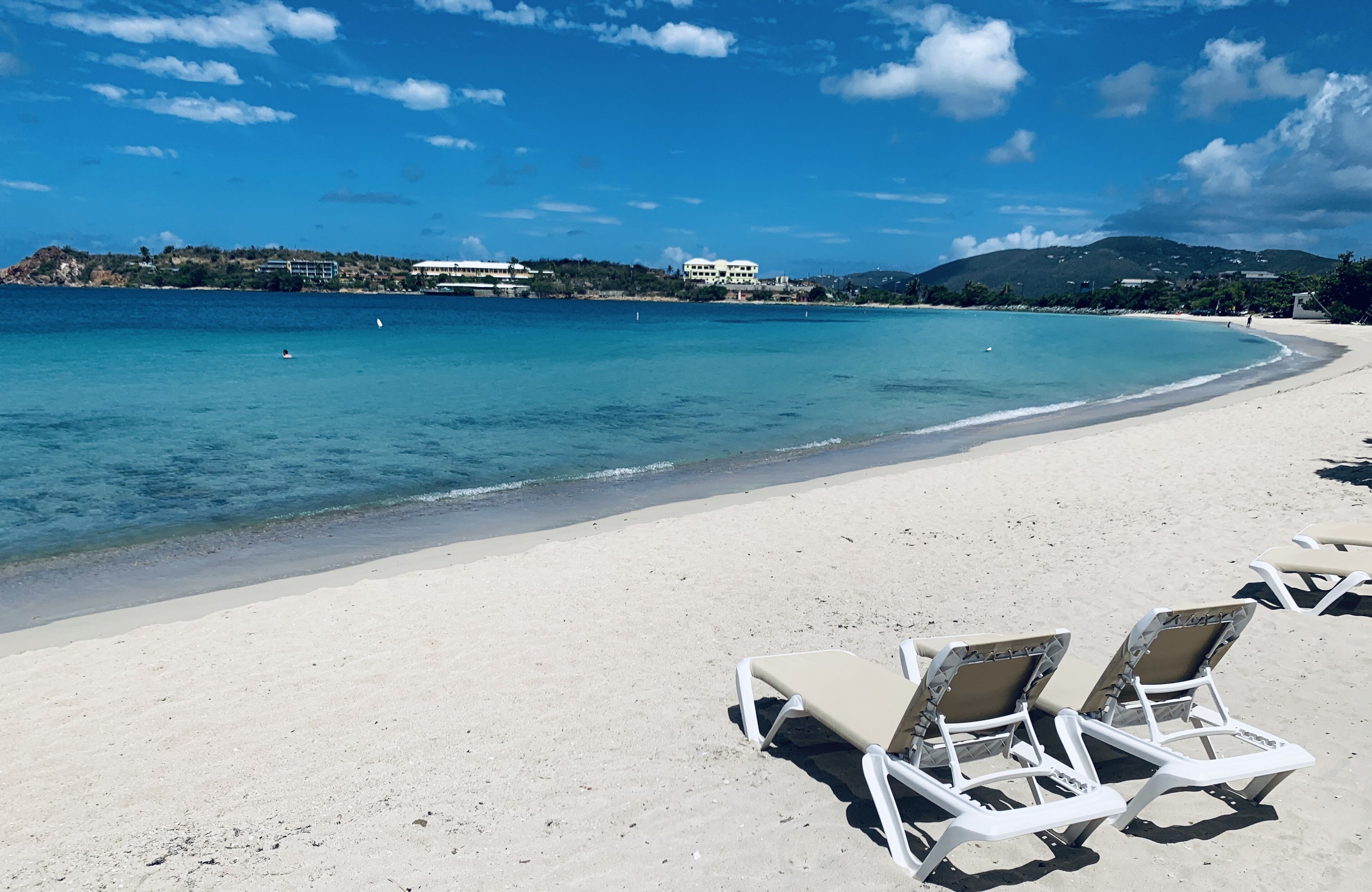 Una playa con sillas reclinables blancas y una vista despejada del mar y edificios lejanos.
