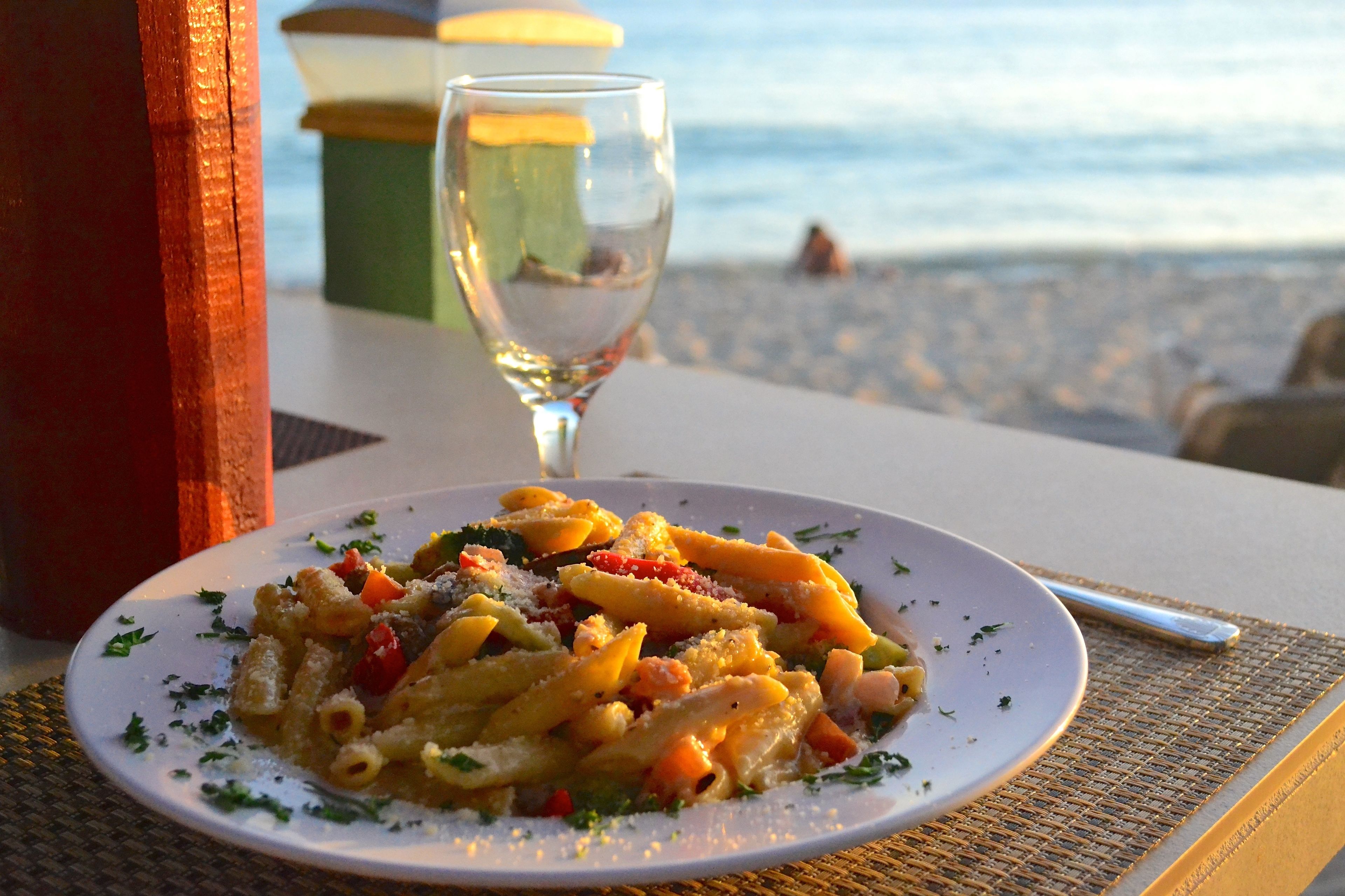 A plate of pasta with vegetables and cheese, a glass of water, and a view of the beach in the background.