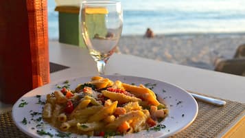 Une assiette de pâtes aux légumes et au fromage, un verre d’eau et, en arrière-plan, une vue sur la plage.