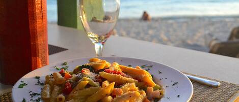 A plate of pasta with vegetables and cheese, a glass of water, and a view of the beach in the background.