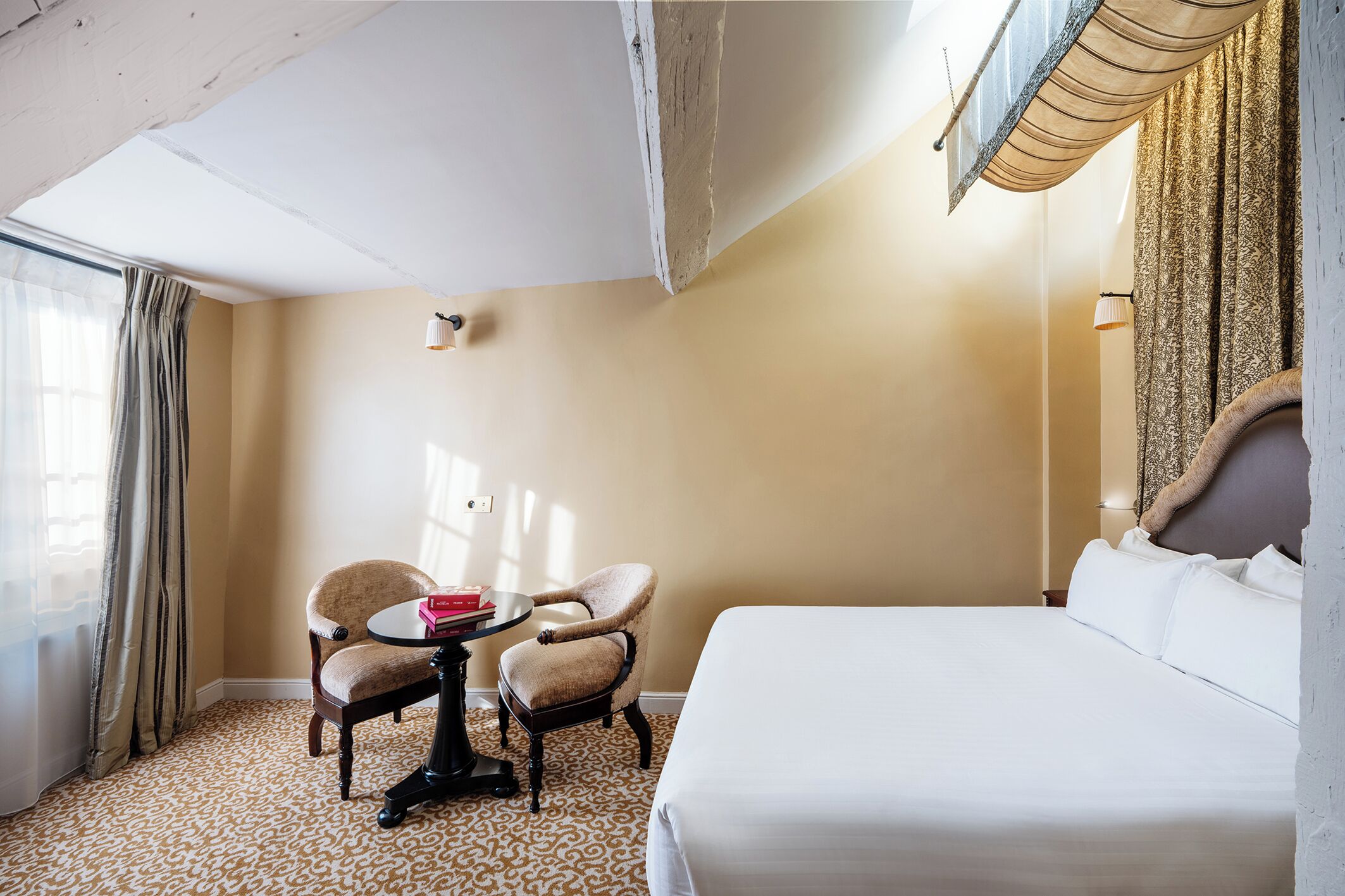 A hotel room with a bed, two chairs, a small table, a lamp, and a window with curtains.