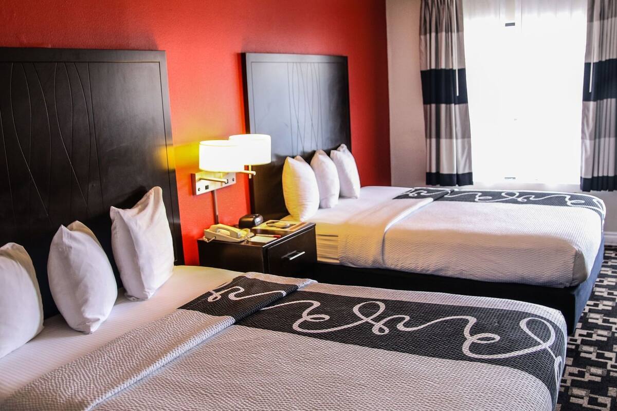 A hotel room with two beds, a nightstand, a lamp, and a view of the outdoors.