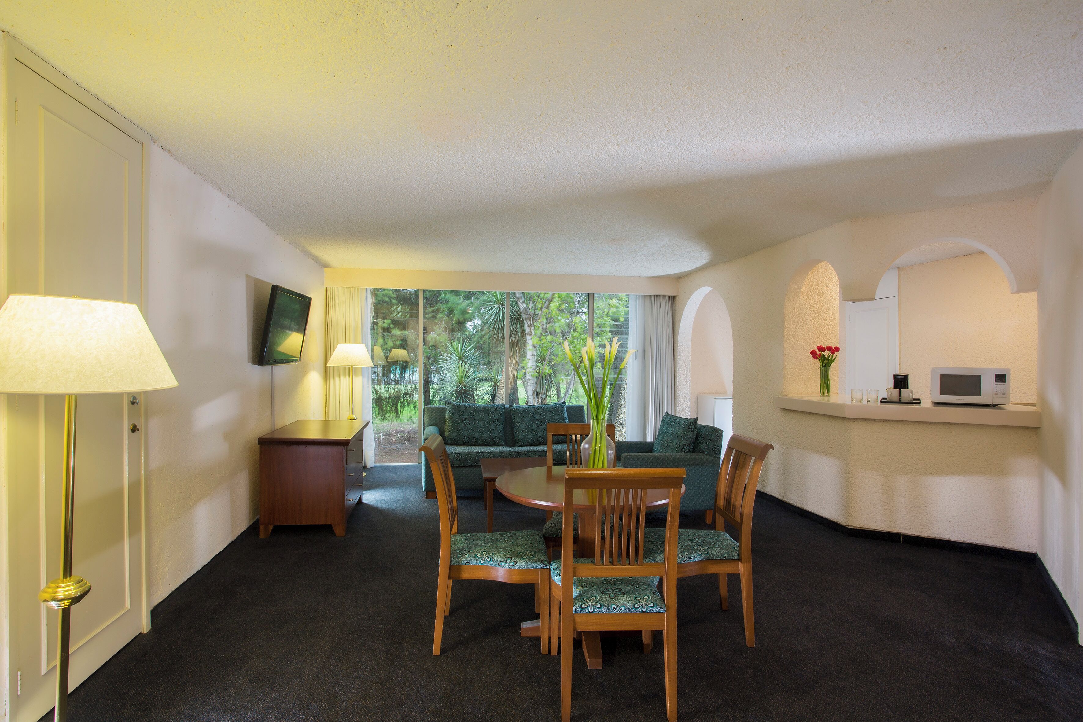 Habitación de hotel con comedor que incluye una mesa redonda y cuatro sillas, un sofá, un televisor montado en la pared y una pequeña cocina con microondas.