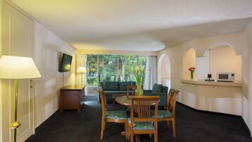 Habitación de hotel con comedor que incluye una mesa redonda y cuatro sillas, un sofá, un televisor montado en la pared y una pequeña cocina con microondas.