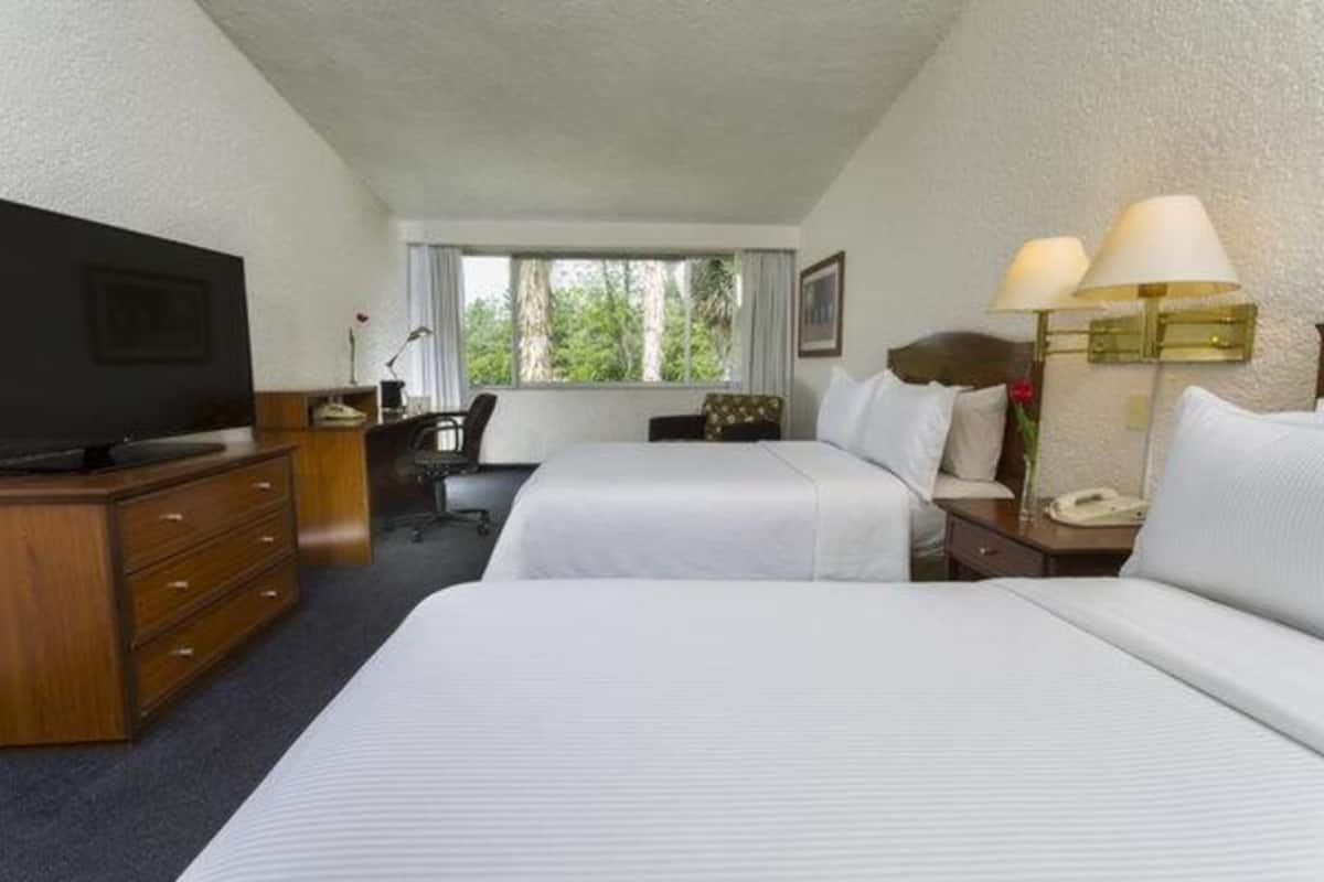 Ein Hotelzimmer mit zwei Betten, einem Schreibtisch mit Fernseher, einer Lampe und einem Fenster mit Blick auf Bäume.