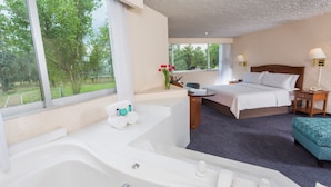 Habitación de hotel con un ventanal grande, una bañera, una cama, una mesita de noche con una lámpara y una pequeña mesa con un florero.