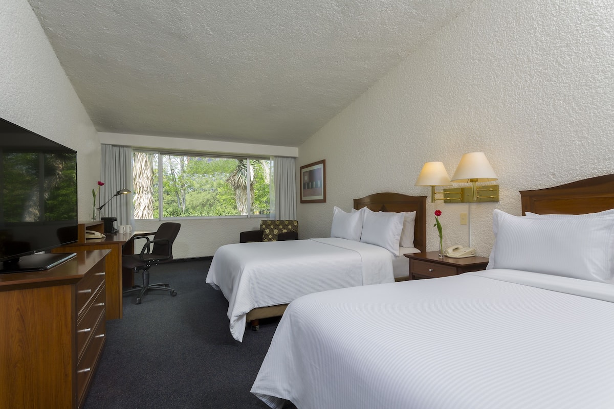 Ein Hotelzimmer mit zwei Betten, einem Schreibtisch mit Fernseher, einem Sessel, einem Fenster mit Vorhängen und einer Lampe.