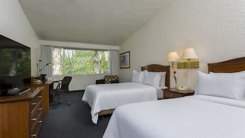Habitación de hotel con dos camas, un escritorio con televisor, una silla, un ventanal con cortinas y una mesita de noche con lámparas.