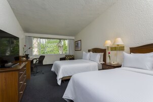 A hotel room with two beds, a desk with a TV, a chair, a window with curtains, and a nightstand with lamps.