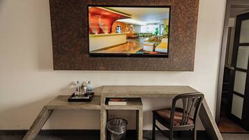 Habitación de hotel con televisión en pared texturizada, escritorio de madera con silla, bote de basura y una mesita con un libro y una botella.