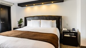 A hotel room with a large bed, a bedside table with a phone and a digital clock, and a wall-mounted air conditioning unit. - Riande Urban Hotel (Panama City)