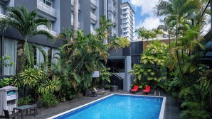 A swimming pool with lounge chairs and a covered seating area, surrounded by lush greenery and modern buildings. - Riande Urban Hotel (Panama City)