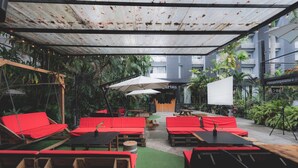 An outdoor seating area with red cushioned benches and tables, surrounded by greenery and a corrugated metal roof. - Riande Urban Hotel (Panama City)