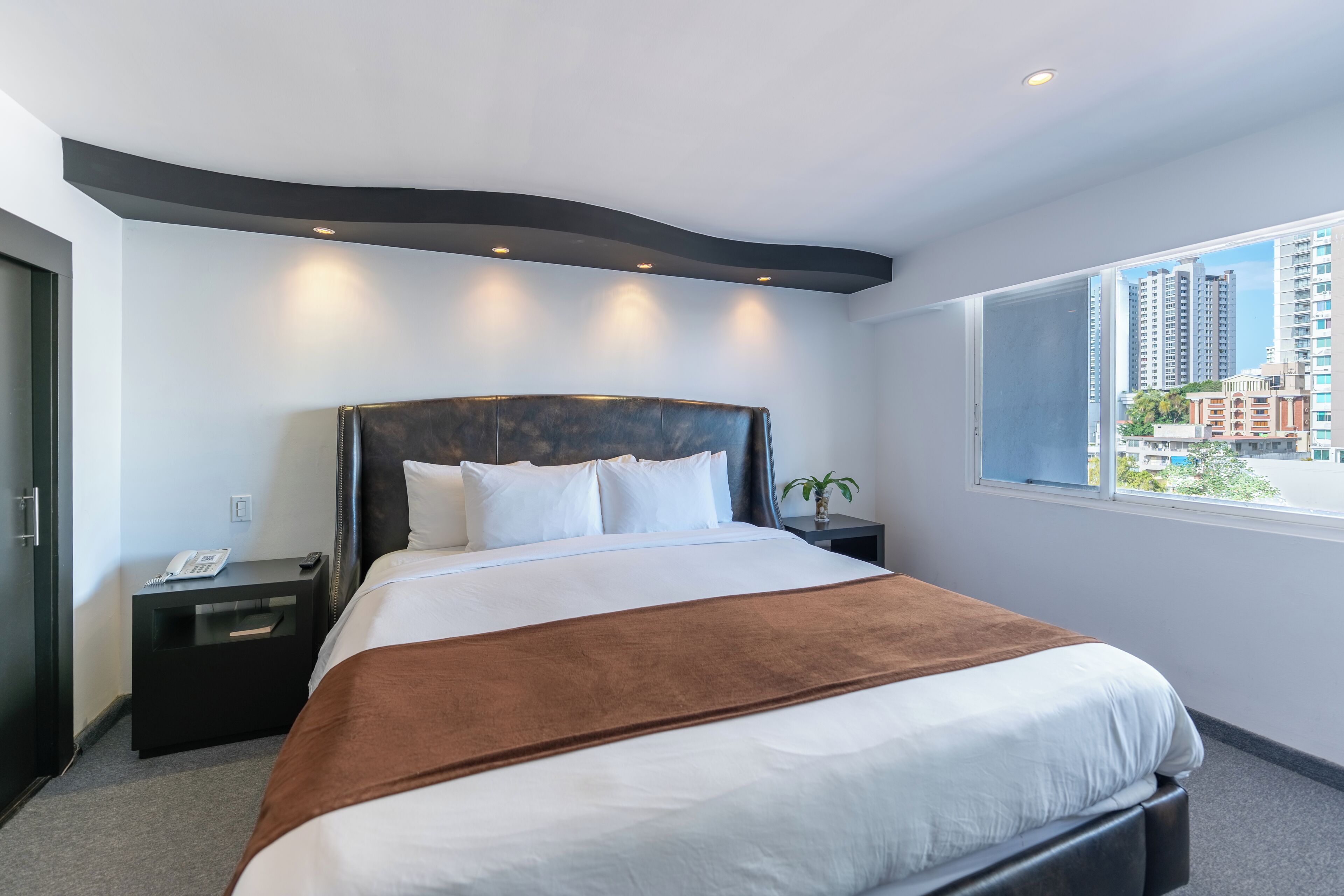 A hotel room with a large bed, a bedside table with a phone, a window with a city view, and a dark ceiling.