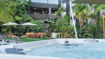 A hot tub surrounded by lounge chairs and umbrellas, with a pool and greenery in the background.