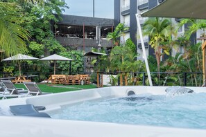 A hot tub surrounded by lounge chairs and umbrellas, with a pool and greenery in the background.