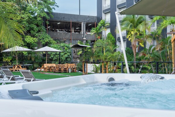 Un bain à remous entouré de chaises longues et de parasols, avec une piscine et de la verdure en arrière-plan.
